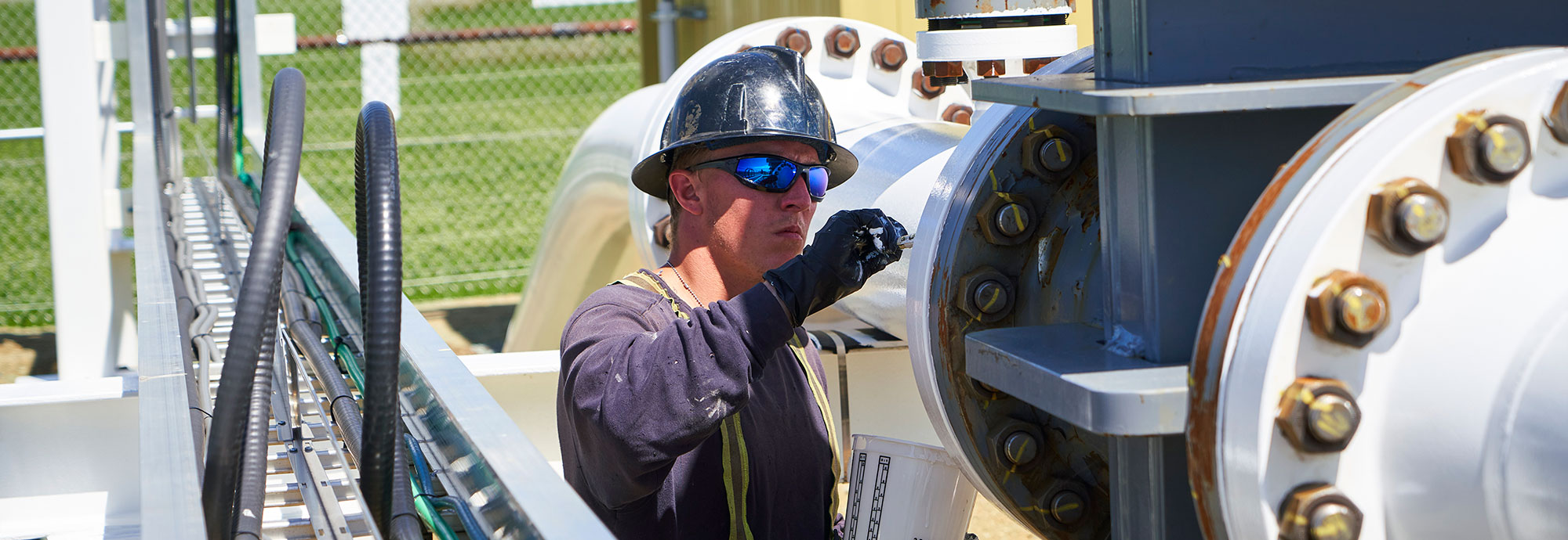 Un homme portant un chapeau de sécurité, des lunettes de soleil, des gants de travail et une chemise à haute visibilité peignant un segment de pipeline en surface avec de la peinture blanche.