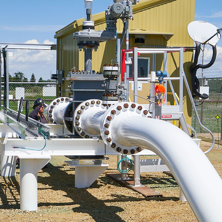 Un segment de pipeline, peint en blanc, vient au-dessus du sol à une station de pompage, avec un bâtiment jaune derrière le segment à l'intérieur de la station clôturée.