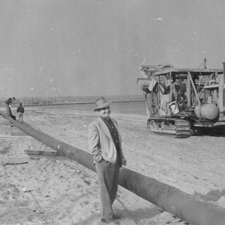 Image en noir et blanc d'un homme dans un manteau de costume et un chapeau debout à côté d'un segment de pipeline, avec et vieille excavatrice à proximité.