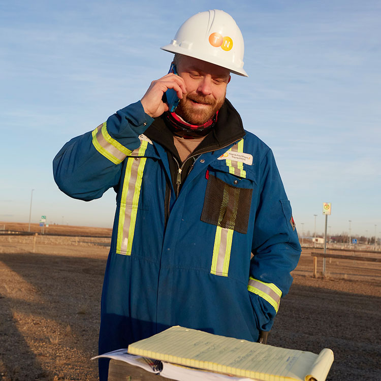 Un technicien de pipeline PTNI dans sa combinaison bleue et son chapeau de sécurité parle sur un téléphone portable.