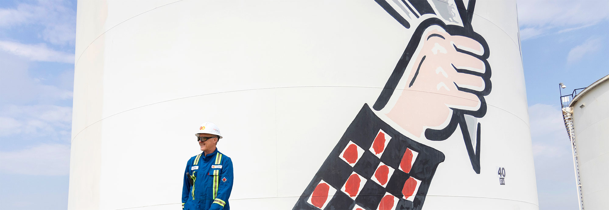 Un employé de PTNI portant une combinaison bleue, un casque de sécurité et des lunettes de sécurité marche devant un réservoir de stockage de carburant à Calgary.